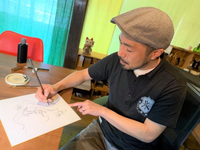 木の机に向かって集中して筆を動かし、繊細な線画を描いている帽子姿の小山康博さんの写真