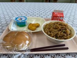 ピンクのトレーに給食が左から時計回りにヨーグルト、チキンナゲット、牛乳、焼きそば、ミルクパンが並んでいる写真