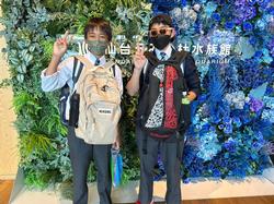 緑色の植物と、青色の植物で飾られた水族館の看板の前で2人の男子生徒がピースサインをして立っている記念写真