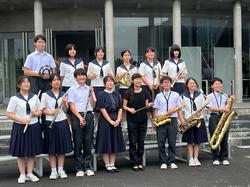 2人の女性を中央にして、複数の男女の生徒が、制服姿でフルートやクラリネット、サックスなどの楽器を持ち、集合写真を撮影している様子の写真