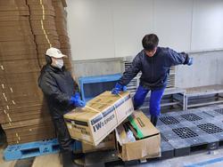 倉庫のような場所で、作業着を着た男性とジャージ姿の男子生徒が、段ボールを協力して持ち上げている様子の写真