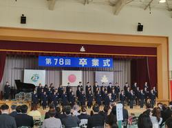 たくさんの保護者らが席に座る中、体育館の舞台前に置かれたひな壇に、制服を着た大勢の生徒たちが立ち、ステージには「第78回 卒業式」と書かれた横断幕が掲げられている写真