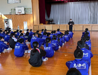 体育館で、青いジャージを着た大勢の生徒たちが床に座り、ステージ前に関係者が立ち話をしている不審者対応訓練の様子の写真