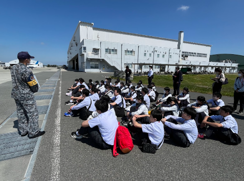 格納庫のような白い大きな建物の前の舗装された広場で、迷彩服と制帽を着用し「教育隊」の腕章をした男性が説明をし、制服姿の生徒達が体育座りをして聞いている写真