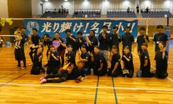 体育館でスポーツウェアを着た複数の人々が、「光り輝け!名足小!!」と書かれた横断幕の前で、笑顔でピースサインをしながら集合している写真