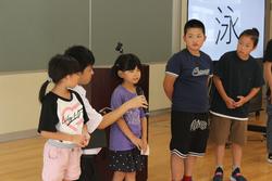 室内で、教壇の前に膝たちしている男性教師がマイクを持って横に立つ女の子に話しかけている様子の写真