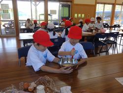 赤い帽子をかぶった2人の生徒が、ひとりはボウルを持ち、もうひとりがかき混ぜるなど協力しながらお菓子作りを体験している様子の写真