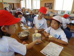 赤白帽をかぶった生徒たちがテーブルを囲んで座り、アイスクリームメーカーや金属のボウルが置かれ、小さなカップに次ぎ分け、カップに入ったアイスクリームの試食をしている様子音写真