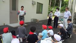 建物前の広場で、複数の生徒たちが地面に座り、生徒たちの前に立っている男性の話を聞いている社会科見学の様子の写真