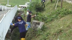 白い橋の横の土手で、複数の保護者の方が鎌や手を使って雑草や木を刈り取り、斜面の清掃作業を行なっている様子の写真
