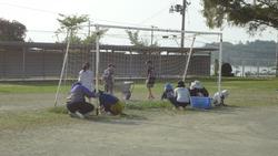 広々とした屋外グラウンドで、保護者の方たちがサッカーゴールを囲み、草むしりや清掃活動に取り組んでいる様子の写真