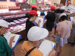 見学に訪れた生徒の皆さんが、店内の陳列棚の前で、店員の男性から説明を受けながら、クリップボードにメモを取っている様子を写した写真。