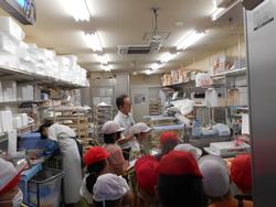 業務用の棚や調理器具が並ぶバックヤードを生徒の皆さんが見学しており、店員の男性の説明を聞いている様子の写真