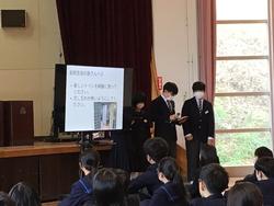 体育館の舞台前に設置した大きなモニターに資料が映し出され、その横に並んで立った3人の生徒たちが、前に並んだ生徒たちへ話をしている写真