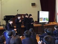 舞台前に設置されたモニターに資料が映し出され、横に並んだ4人の生徒が、前に座った生徒たちに向かって発表をしている写真