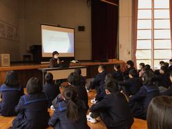 舞台上に置かれたスクリーンに資料が映し出され、その前に座った校長先生の話を、整列して床に座ったたくさんの生徒たちが聞いている様子の写真