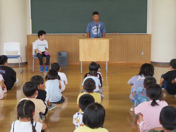 たくさんの児童が床に整列して座っている中、一人の男子児童が教卓に立ち、皆の前で話をし、横の椅子には別の児童が座っている始業式の写真