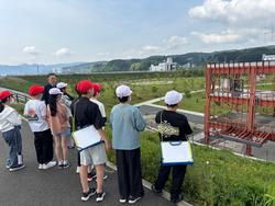 赤や白の帽子をかぶった小学生たちが、屋外でクリップボードを手にしながら並び、鉄骨だけが残った防災庁舎を見学している写真