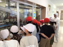 ガラス越しに、赤と白の帽子をかぶった複数の子供たちが、全身白の作業着を着た作業員の説明を聞きながら機械が稼働している様子を見学している写真