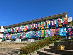 青空の下、学校の校舎の二階と三階に、多様な色とデザインの縦長の旗がずらりと並べて飾られている様子の写真