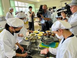 2台のカメラによる撮影が行われる中、白い調理服と帽子、マスクを身に着けた数名の生徒たちが調理台を囲んで、料理の盛り付けや洗い物をしている様子の写真