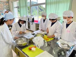 白い調理服と帽子、マスクを身に着けた4人の生徒が、白衣と三角巾を身に着けた大学生と調理台を囲み、野菜の下処理をしている様子の写真