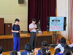 舞台の前に置かれた大きなモニターに資料が映っており、その横に並んで立った男女2人の生徒が発表を行っているのを、その前に並んで座った生徒たちが聞いている様子の写真