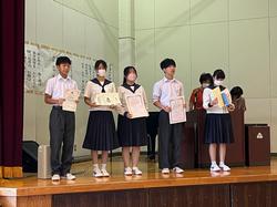 壇上で制服を着た5人の生徒が、賞状などを手にして横一列に並んで立っている写真