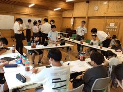 木製の壁の部屋で、複数の学生たちが机に座って議論をしており、先生らしき大人が生徒たちの間を歩きながら指導している様子の写真