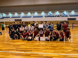 アリーナの屋内コートで、バレーのユニフォームを着た多数のPTAの保護の方々が集合し、笑顔で写っている集合写真