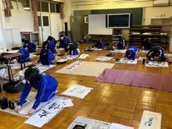 広い教室の床に新聞紙や敷物を広げ、ジャージを着た複数の生徒たちが書道道具を使って大きな紙に文字を書いている写真