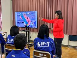 教室で赤色のジャージを着た女性が、椅子に座った生徒たちにスクリーンを指差し、映像を見せながら説明している写真