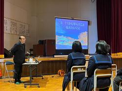 舞台の上に置かれたスクリーンに資料が映し出され、その横でマイクに向かって立った菅原さんが、椅子に座った生徒たちの方を向いて話をしている写真