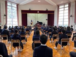 校章旗が飾られた舞台の方を向き、整然と並んだ椅子に座った大勢の生徒たちを後方から写した写真