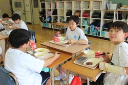 教室の中で小学生の子どもたちが給食を食べており、机を囲んで座りながら、それぞれのトレイにのった食事を楽しんでいる様子の写真