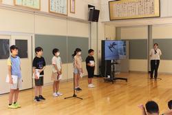 広い室内で5人の生徒が本を手に持ち横一列に並んでいる写真