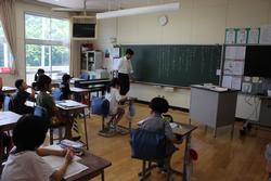 生徒たちが教室の机に座り、男性の先生が黒板の前に立って授業を進行している様子の写真