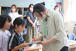 子供たちが何かを提出し、それを先生が受け取って記入している、学習活動の一環の様子の写真