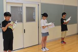 広い室内で、三人の小学生が間隔をあけて立ち、それぞれ本を手に持ち真剣に朗読をしている様子を写した写真
