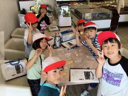 帽子をかぶった複数の子どもたちが、店内のテーブルを囲み、ピースサインをしたり、何かを食べたりしながら笑顔で集合写真を撮っている様子の写真