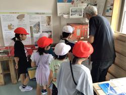 帽子をかぶった複数の子どもたちが、白髪の男性に何かを教わりながら、展示室のパネルや資料を熱心に見学している様子の写真