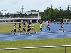 青いトラックと芝生の広がる競技場で、緑や黒のユニフォームを着用した複数の女子選手たちが、レースをしている様子の写真