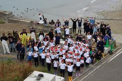 大勢の子どもたちと大人たちが海辺の護岸で、白と赤の帽子をかぶった子どもたちは前列に、大人は後列に分かれてカメラに向かって手を振り集合している様子の写真