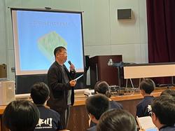 黒い服を着た男性がマイクを持ち、プロジェクターに映し出された図形を背景に講演をしており、その話を聞く学生たちの後姿が写し出された写真