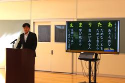 スーツ姿の先生が、演台にあるマイクに向かってスピーチをしており、横には「あたりまえ」などの言葉が書かれたスクリーンが設置されている様子の写真