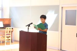 ホワイトボード前にある演台に立つ小学生の男の子が、緑色の本を手に持ちマイクに向かって1学期の振り返りをを発表している様子の写真