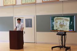 演台のマイクに向かって話をする男性と、隣のモニターに「やってみよう!」と書かれた画面が映し出されている始業式の様子の写真