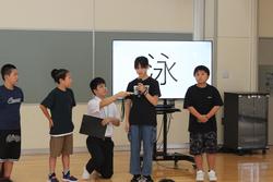 後方のモニターには大きく「泳」という漢字が映し出されている前に4人の生徒が立ち、そのうちの一人の生徒に男性教師がマイクを向けている写真