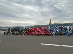 海を挟んだ低い山並みの風景を背景に、舗装された道路脇に緑、赤、水色の法被を着た大勢の人々がチームに分かれて整列して撮影した集合写真