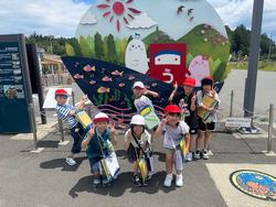 キャラクターが描かれた大きな看板の前で、複数の小学生たちが赤白帽をかぶり、ピースサインをして集合している写真